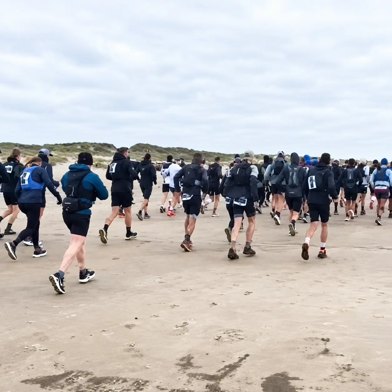 Runners with race bibs and packs setting off across a wide sandy beach