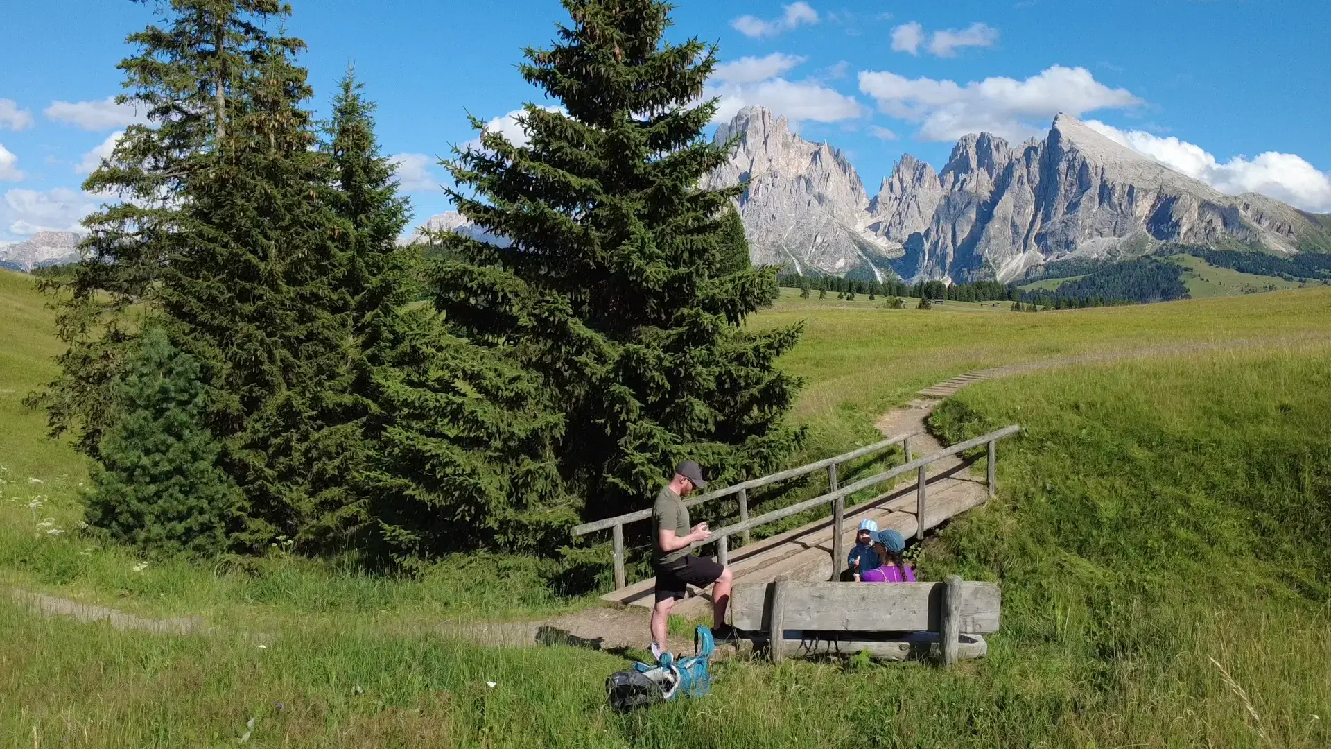 A family break on a hike, by a wooden bridge beneath jagged mountain peaks