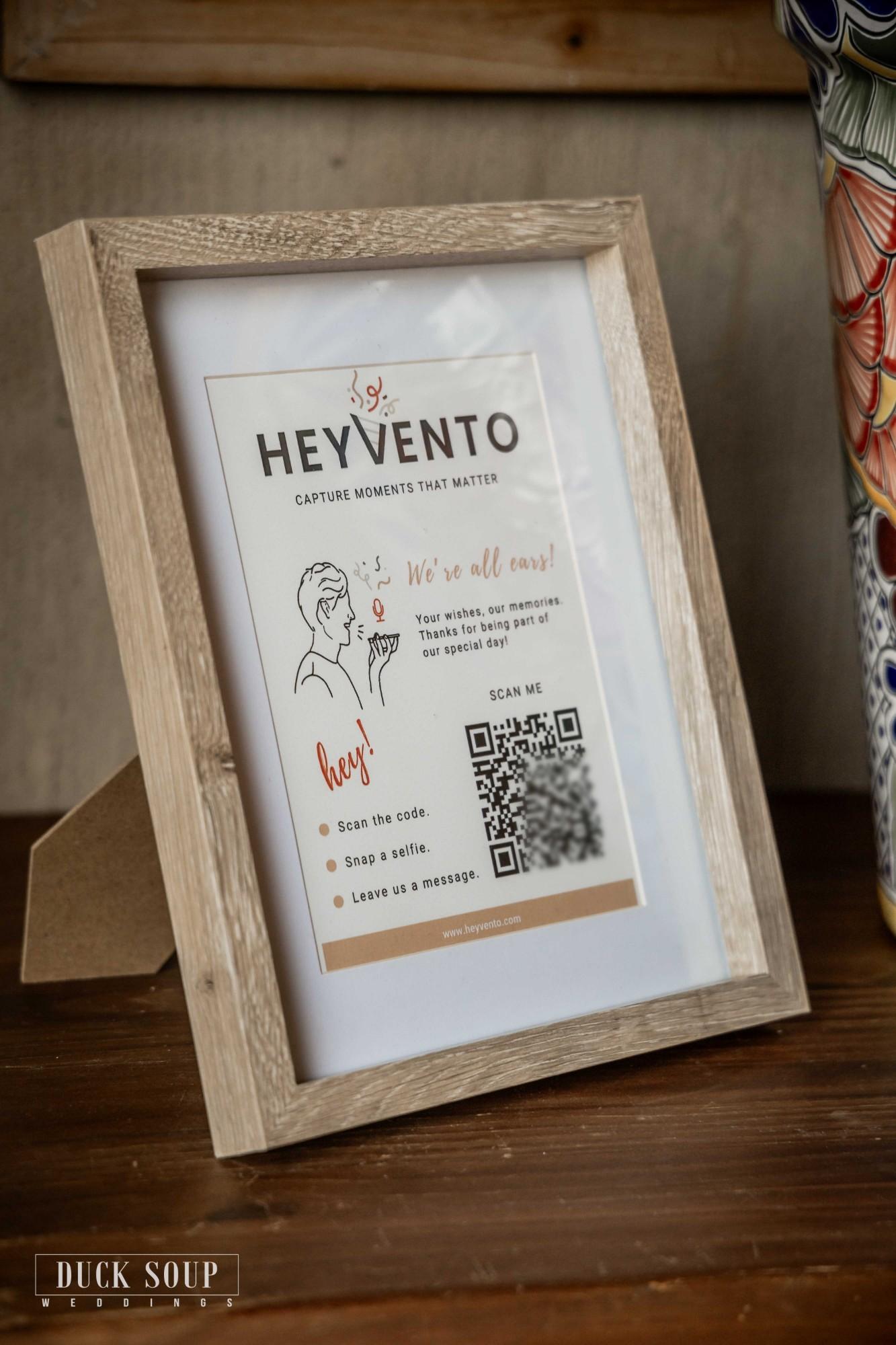 A framed Heyvento sign on a wedding table: scan the code, snap a selfie, leave us a message